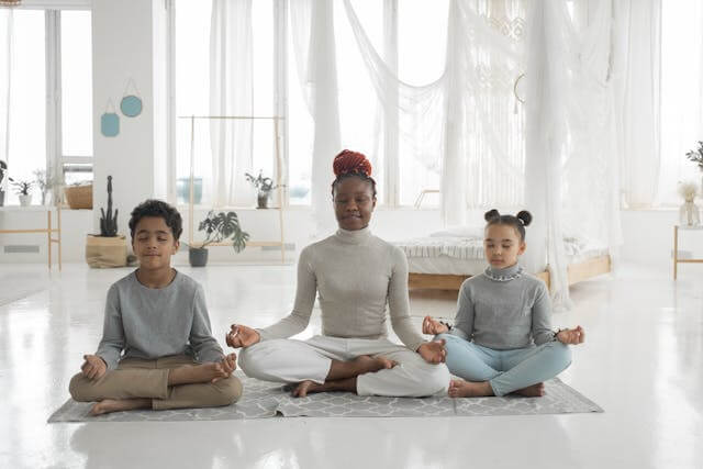 yoga teacher and 2 children in lotus pose on a rug in home